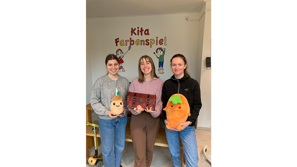 Drei Studentinnen lächeln in die Kamera und halten ein Kartoffel- und ein Karotten-Kuscheltier sowie eine Schatztruhe in den Händen.